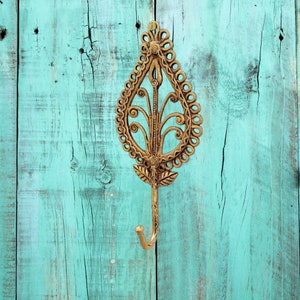 May include: Ornate brass wall hook with a teardrop-shaped design. The hook features a decorative openwork pattern and a curved hook at the bottom. The hook is mounted on a distressed turquoise wooden surface.
