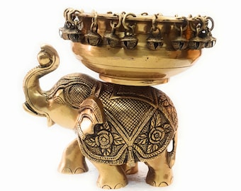 May include: A gold metal elephant figurine with intricate details, holding a round bowl with a decorative border of small bells.