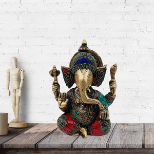May include: A gold-colored brass statue of Ganesha, the Hindu god of beginnings, wisdom, and prosperity. The statue is decorated with colorful gemstones and intricate details. Ganesha is depicted sitting with his trunk raised and holding a sweet in his hand.