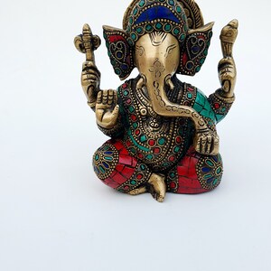 May include: A gold-colored Ganesha statue with intricate details and colorful gemstone accents. The statue is sitting with its legs crossed and its trunk curled up.