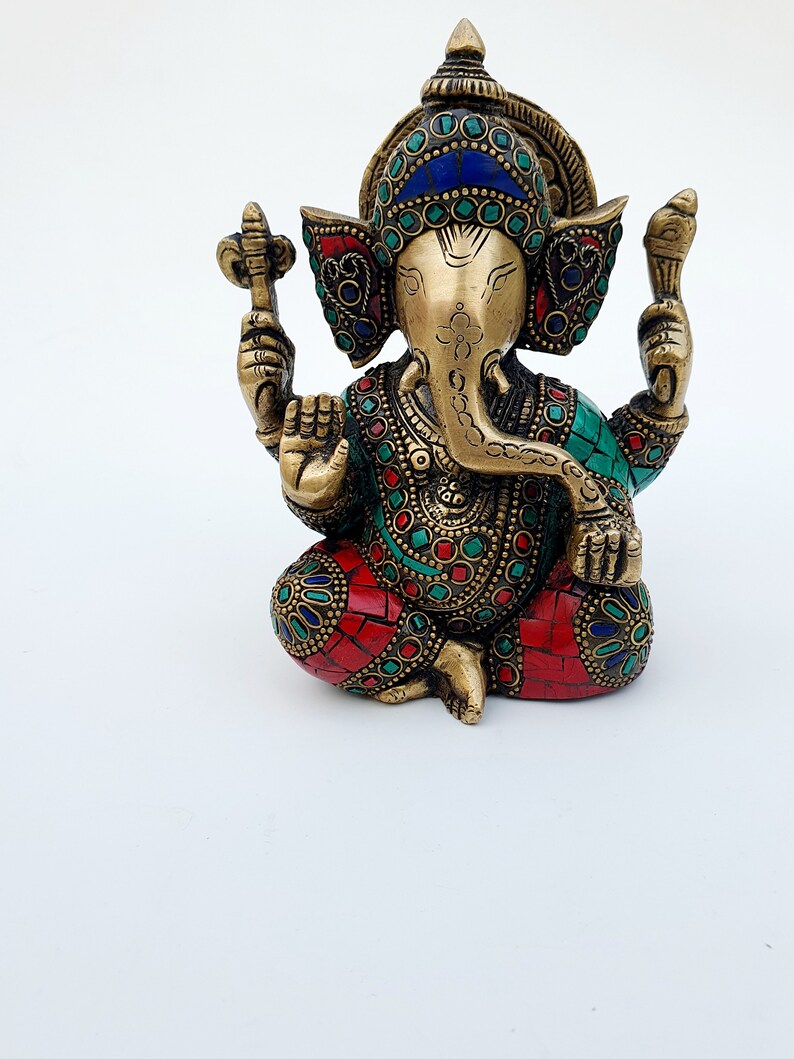 May include: A gold-colored Ganesha statue with intricate details and colorful gemstone accents. The statue is sitting with its legs crossed and its trunk curled up.