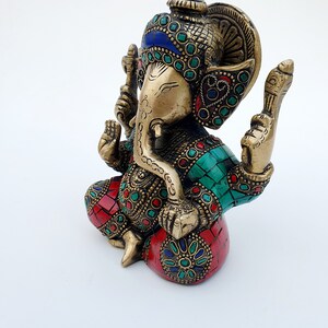 May include: A gold-colored brass statue of Ganesha, the Hindu god of wisdom and prosperity. The statue is decorated with colorful gemstones and intricate details. The statue is sitting on a base with a red and green mosaic design.