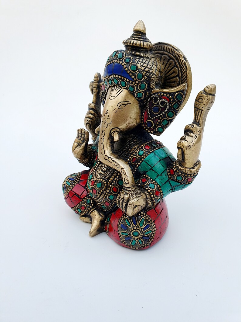 May include: A gold-colored brass statue of Ganesha, the Hindu god of wisdom and prosperity. The statue is decorated with colorful gemstones and intricate details. The statue is sitting on a base with a red and green mosaic design.
