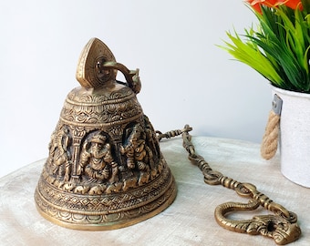 Könnte beinhalten: Eine goldfarbene Metallglocke mit aufwendigen Schnitzereien von Gottheiten und Fabelwesen. Die Glocke hängt an einer Kette mit einem dekorativen Haken.