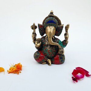 May include: A small, ornate brass statue of Ganesha, the Hindu god of wisdom and prosperity. The statue is decorated with colorful gemstones and intricate details. The statue is sitting on a white surface.