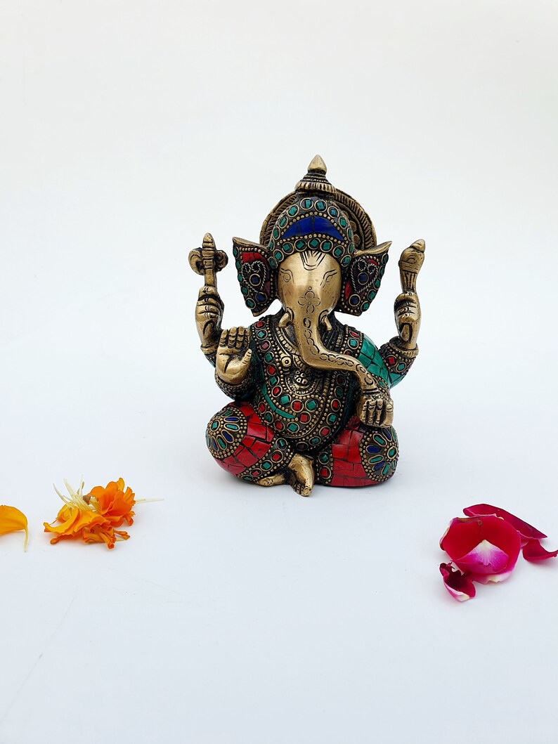 May include: A small, ornate brass statue of Ganesha, the Hindu god of wisdom and prosperity. The statue is decorated with colorful gemstones and intricate details. The statue is sitting on a white surface.