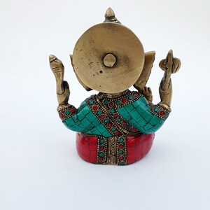 May include: A gold-colored brass statue of Ganesha, the Hindu god of wisdom and prosperity, with red and green gemstone accents. The statue is sitting with its back to the viewer, and its trunk is raised.