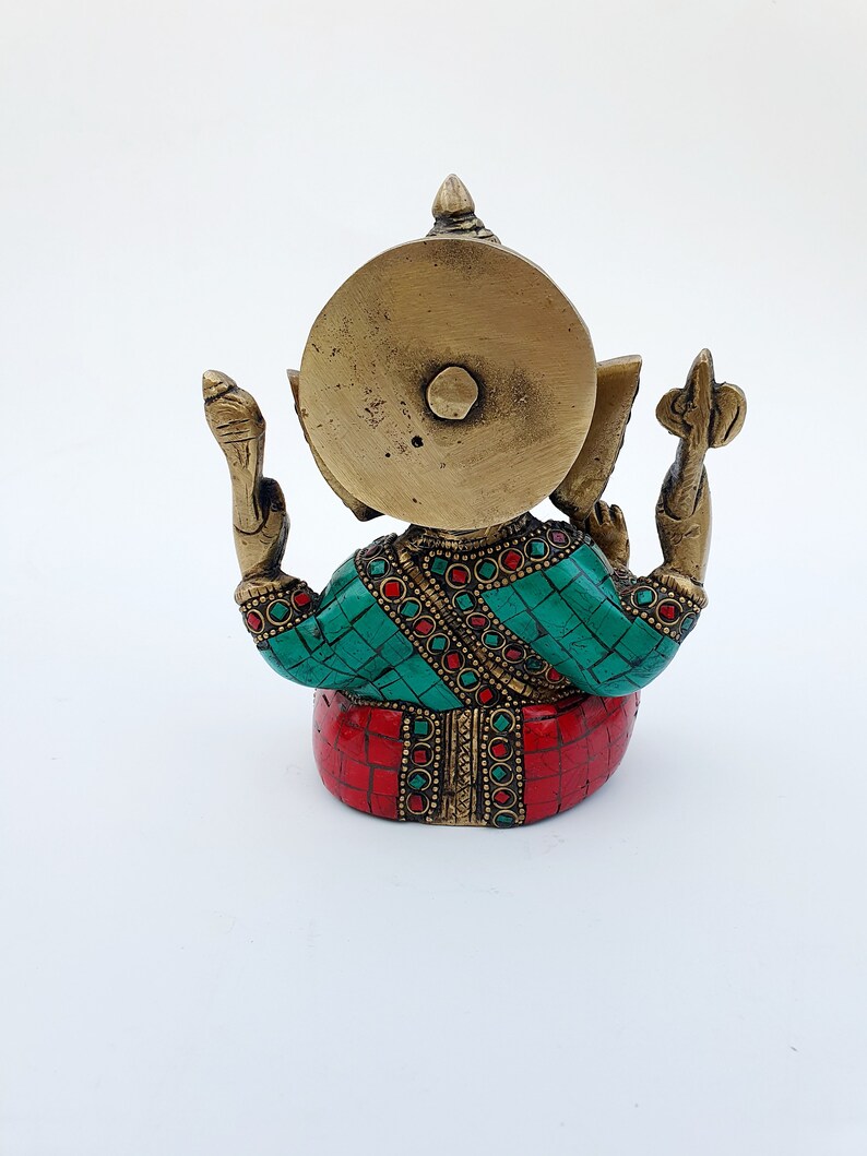 May include: A gold-colored brass statue of Ganesha, the Hindu god of wisdom and prosperity, with red and green gemstone accents. The statue is sitting with its back to the viewer, and its trunk is raised.
