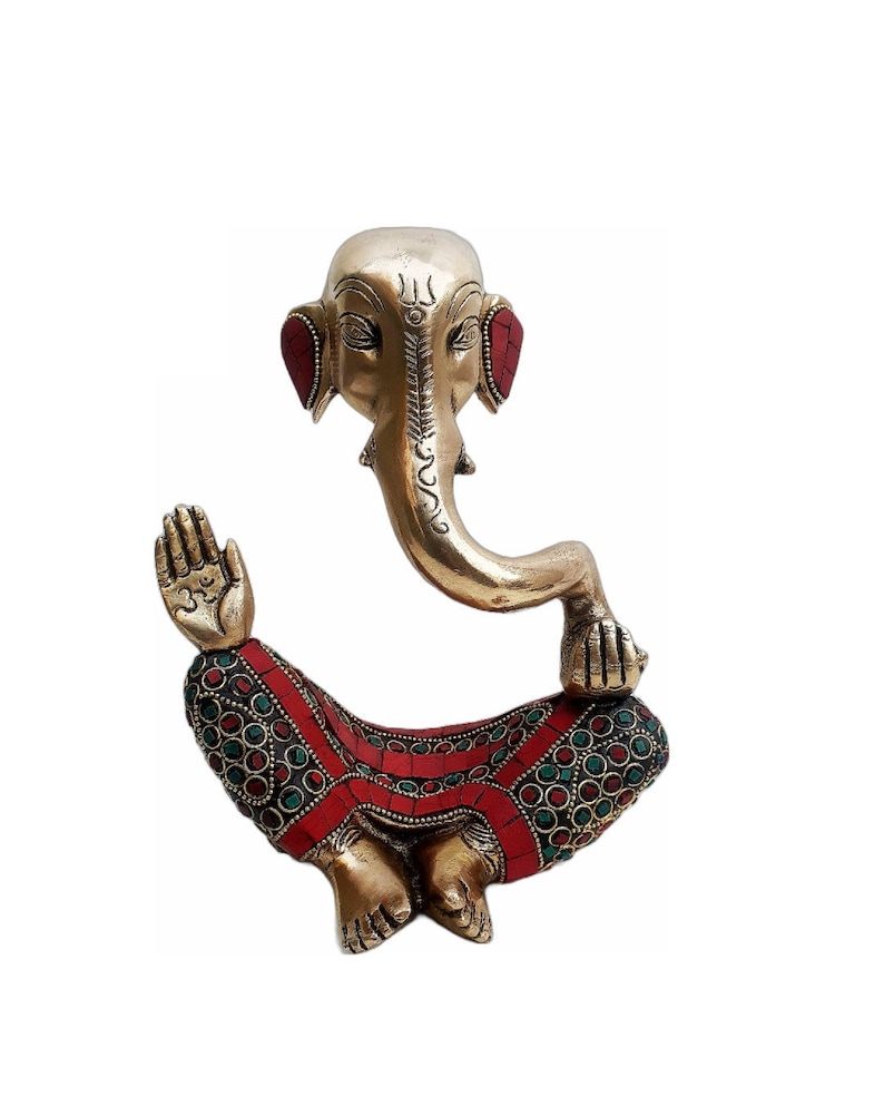 May include: A gold-colored brass statue of Ganesha, the Hindu god of wisdom and prosperity, sitting in a meditative pose. The statue is decorated with red and green gemstones.