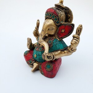 May include: A gold-colored brass statue of Ganesha, the Hindu god of wisdom and prosperity. The statue is decorated with red and green gemstones and has a detailed design.