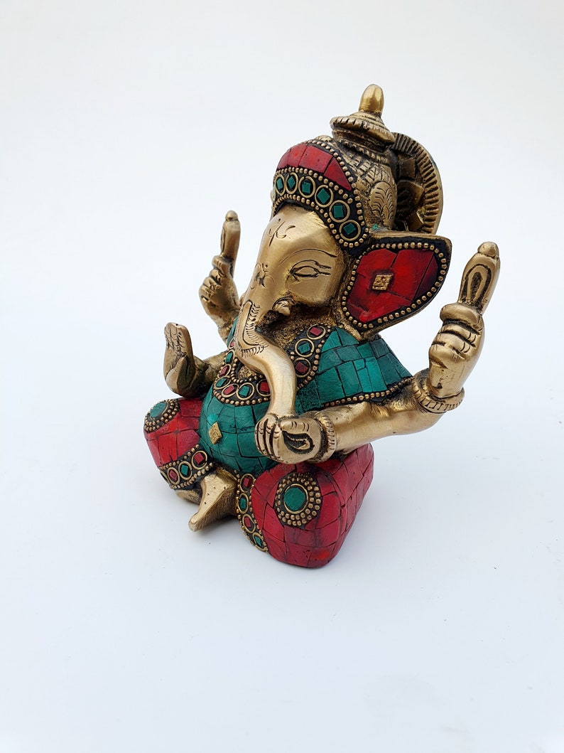 May include: A gold-colored brass statue of Ganesha, the Hindu god of wisdom and prosperity. The statue is decorated with red and green gemstones and has a detailed design.