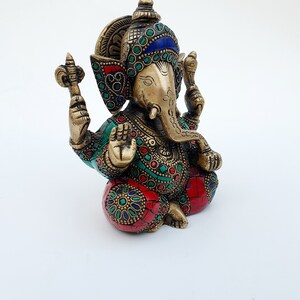 May include: A brass statue of Ganesha, the Hindu god of wisdom and prosperity, with intricate details and colorful gemstones. The statue is sitting with its legs crossed and its trunk curled up. The statue is decorated with red, green, and blue gemstones.