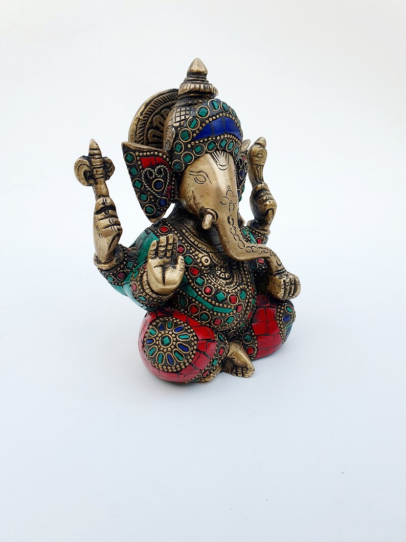 May include: A brass statue of Ganesha, the Hindu god of wisdom and prosperity, with intricate details and colorful gemstones. The statue is sitting with its legs crossed and its trunk curled up. The statue is decorated with red, green, and blue gemstones.