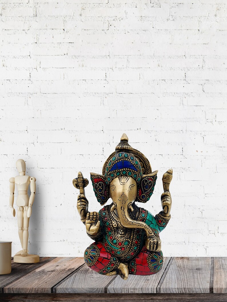 May include: A gold-colored brass statue of Ganesha, the Hindu god of beginnings, wisdom, and prosperity. The statue is decorated with colorful gemstones and intricate details. Ganesha is depicted sitting with his trunk raised and holding a sweet in his hand.