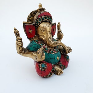 May include: A gold-colored brass statue of Ganesha, the Hindu god of beginnings, with red and green gemstone accents. The statue is sitting with its legs crossed and its hands in a mudra.