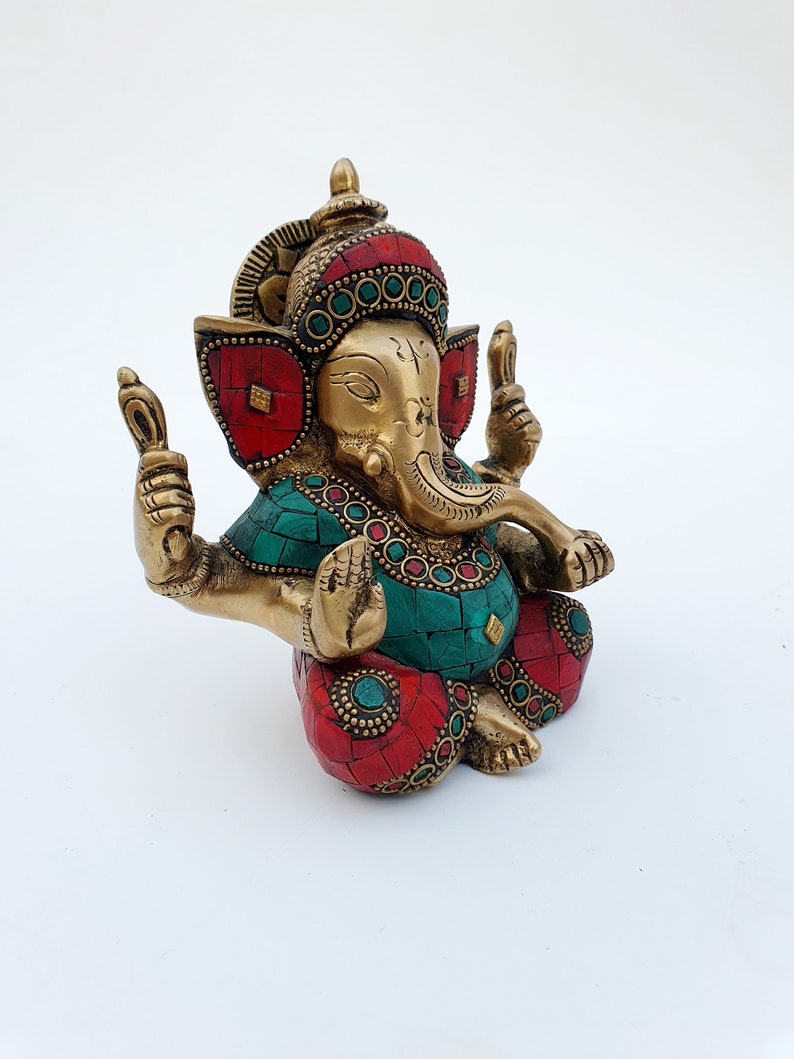 May include: A gold-colored brass statue of Ganesha, the Hindu god of beginnings, with red and green gemstone accents. The statue is sitting with its legs crossed and its hands in a mudra.
