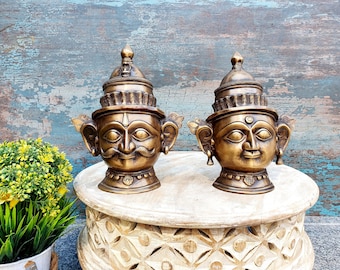 May include: Two ornate brass sculptures of human heads with intricate details. The sculptures are placed on a wooden pedestal with a carved design.