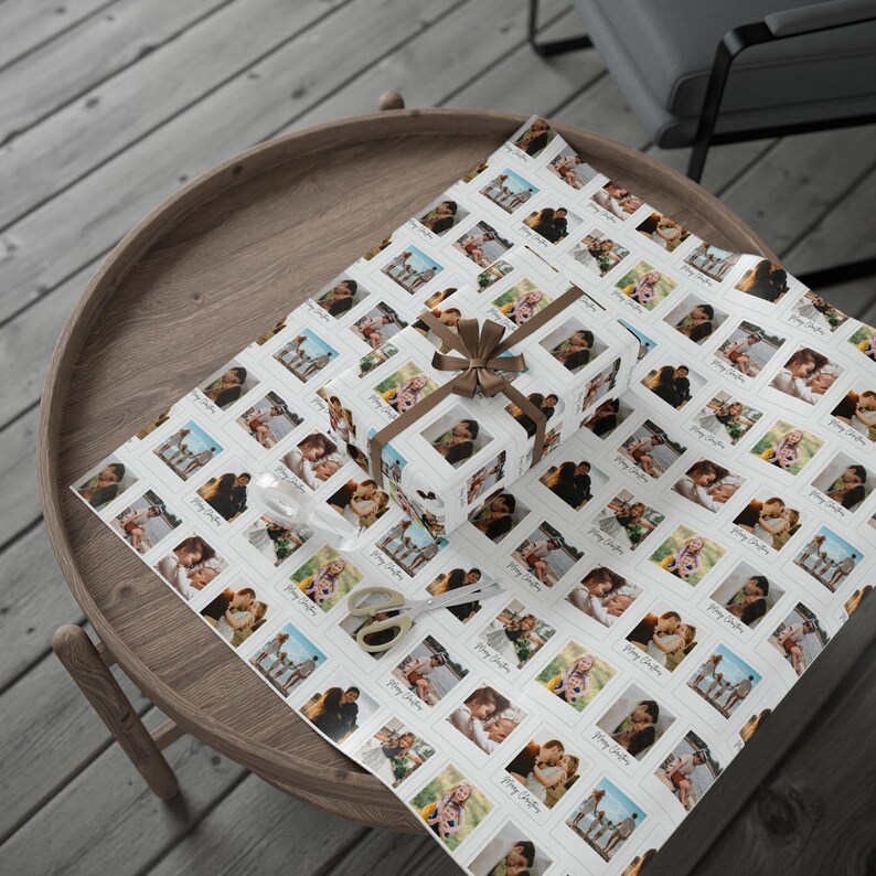 May include: White wrapping paper and a gift box covered in a pattern of framed photographs. The gift box is tied with a brown ribbon. Scissors and a clear ribbon are also on the paper, which is on a wooden table.