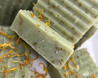 May include: Close-up of several bars of green soap with flecks of dried flowers. The soap is arranged in a pile on a white surface.