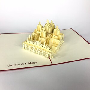 Puede incluir: Una tarjeta de papel pop-up de color crema que presenta un modelo 3D de la Bas&iacute;lica de San Marcos en Venecia, Italia. La tarjeta est&aacute; abierta y el modelo est&aacute; completamente extendido. La tarjeta tiene un borde rojo.