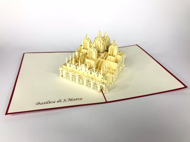 Puede incluir: Una tarjeta de papel pop-up de color crema que presenta un modelo 3D de la Bas&iacute;lica de San Marcos en Venecia, Italia. La tarjeta est&aacute; abierta y el modelo est&aacute; completamente extendido. La tarjeta tiene un borde rojo.