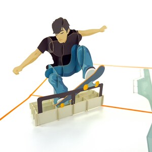 May include: A pop-up card featuring a person skateboarding over a ramp made of cardboard. The person is wearing a black shirt and blue jeans. The skateboard is brown with yellow wheels.