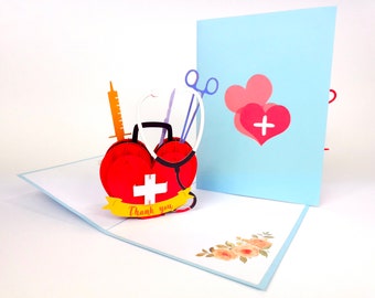 May include: A 3D pop-up card with a red heart-shaped first aid kit with a white cross and a stethoscope. The card has a blue background with a pink heart with a white cross. The card says "Thank you".