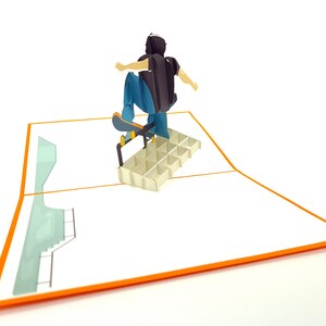 May include: A 3D pop-up card featuring a person skateboarding on a ramp. The card is open and the person is in mid-air, with their skateboard wheels touching the ramp. The card is white with an orange border.
