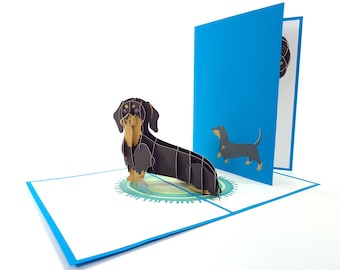May include: A 3D pop-up greeting card featuring a dachshund dog. The card is black and tan, with a blue border and background. The dog is depicted in a detailed, layered paper design, with a smaller illustration on the inside.