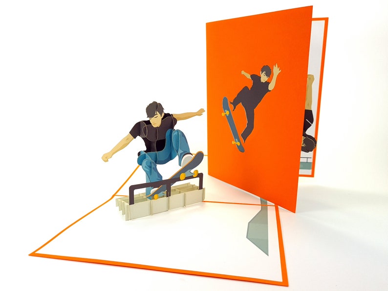 May include: A 3D pop-up card featuring two skateboarders. The card is open, revealing a pop-up of a skateboarder performing a trick on a ramp. The card is orange on the outside and white on the inside.