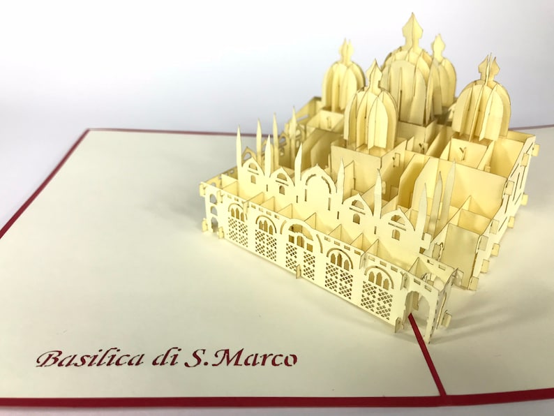 Puede incluir: Una tarjeta de papel pop-up de color crema que representa la Bas&iacute;lica de San Marcos en Venecia, Italia. La tarjeta est&aacute; abierta y se pueden ver los intrincados detalles de la bas&iacute;lica. La tarjeta est&aacute; sobre un fondo rojo y blanco.