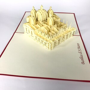 Puede incluir: Una tarjeta de papel pop-up de color crema que representa la Bas&iacute;lica de San Marcos en Venecia, Italia. La tarjeta est&aacute; abierta y se pueden ver los intrincados detalles de la bas&iacute;lica. La tarjeta est&aacute; sobre una superficie blanca.
