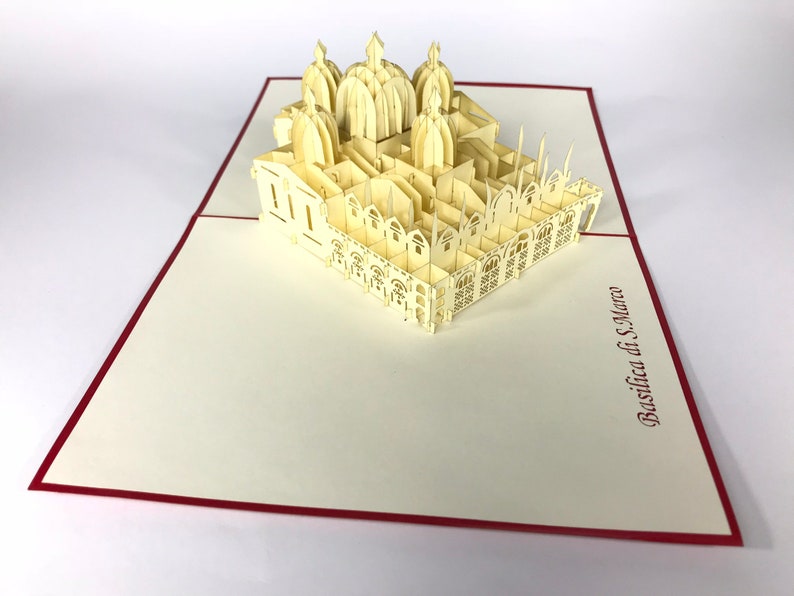 Puede incluir: Una tarjeta de papel pop-up de color crema que representa la Bas&iacute;lica de San Marcos en Venecia, Italia. La tarjeta est&aacute; abierta y se pueden ver los intrincados detalles de la bas&iacute;lica. La tarjeta est&aacute; sobre una superficie blanca.