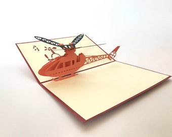 Puede incluir: Una tarjeta de felicitación desplegable con un diseño de helicóptero 3D naranja. La tarjeta abierta revela la aeronave detallada con aspas negras y un interior crema. La tarjeta tiene un borde rojo.