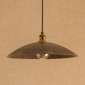 May include: A bronze-colored, dome-shaped pendant light fixture with a textured surface. The light hangs from a black cord and features a brass-colored fixture and a visible lightbulb. The background is a solid, light brown color.