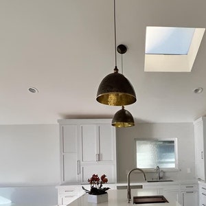 Puede incluir: Una cocina moderna con gabinetes blancos, una isla grande con una encimera blanca y electrodom&eacute;sticos de acero inoxidable. La cocina tiene un tragaluz y dos l&aacute;mparas colgantes doradas sobre la isla.