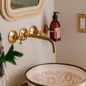 May include: A white bathroom with a vintage-style mirror, gold-toned faucets, and a decorative sink with a pink and white floral pattern. The walls are painted a light beige color, and there are two gold-toned light fixtures with green leaves and gray candles.