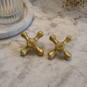 May include: Two polished brass cross-handle faucet knobs with a four-point design. The left knob features the word HOT engraved in black, and the right knob features the word COLD engraved in black, both centered on circular caps.
