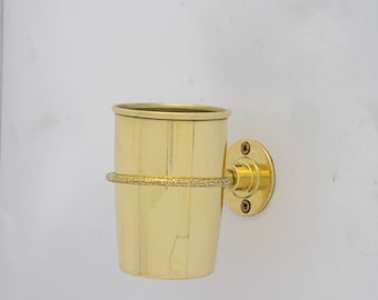 May include: A brass toothbrush holder with a circular wall mount. The holder is cylindrical with a textured band around the middle. The holder is made of shiny, polished brass.