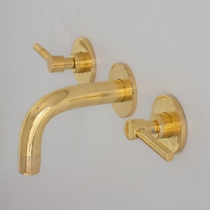 May include: A gold-colored wall-mounted faucet set. The set includes a curved spout and two separate handles, all with a shiny, reflective finish. The faucet is mounted on a white wall.
