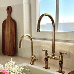May include: Two gold-toned kitchen faucets with a traditional design. The faucets are mounted on a white countertop with white tile backsplash. A wooden cutting board is visible in the background.