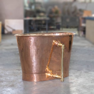 May include: A copper bathtub with a polished finish. The tub has a rounded, oval shape and a brass faucet fixture on the side. The background is a workshop setting with blurred equipment.