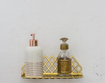 May include: Two gold soap dispensers on a gold metal shelf with a diamond pattern. The soap dispenser on the left is white with gold stripes. The soap dispenser on the right is clear glass with a gold top and a gold design.