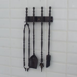 May include: A black fireplace tool set with a wall-mounted holder. The set includes tongs, a shovel, a poker, and a brush. The tools have decorative handles and are made of metal. The set is displayed against a white tiled wall.