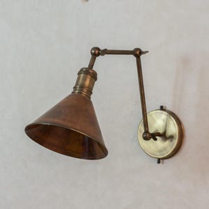 Op de afbeelding: Een wandlamp in vintage stijl met een koperkleurige conische kap en een messingkleurige arm en voet. De verstelbare arm maakt gerichte verlichting mogelijk. Het ontwerp heeft een industriële of retro-esthetiek, geschikt voor binnenverlichting.