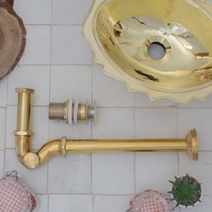 Op de afbeelding: Een verzameling messing badkamerarmaturen, waaronder een wastafelafvoer en een decoratieve wastafel. De wastafel heeft een geschulpte rand en een gehamerde afwerking. De afvoer bestaat uit verschillende onderdelen.