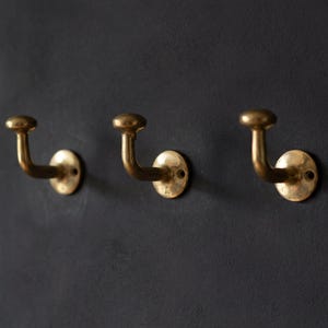 Puede incluir: Tres perchas de latón montadas en una pared gris oscuro. Cada gancho tiene una parte superior redondeada y un brazo curvo que se extiende desde una base circular. Los ganchos están espaciados uniformemente y ofrecen una solución de almacenamiento clásica.