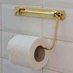 Pode incluir: Um porta-papel higiênico dourado montado em uma parede de azulejos brancos. O suporte tem uma barra horizontal e um braço curvo para segurar o rolo de papel higiênico branco. O design é simples e elegante.