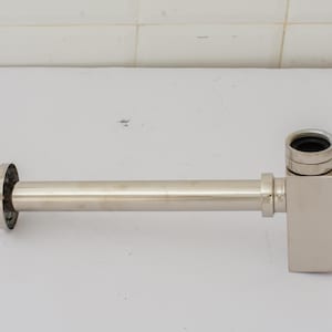 Puede incluir: Un accesorio de fontanería con trampa en P de cromo pulido. El accesorio tiene un cuerpo cilíndrico con una brida circular en un extremo y un componente de forma cuadrada en el otro. La trampa en P está diseñada para evitar que los gases de alcantarillado entren en un edificio.