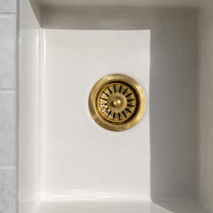 Puede incluir: Un fregadero de cocina rectangular de color blanco con un desagüe circular de metal en tono latón, que presenta un pomo central y ranuras radiales. El fregadero tiene un acabado liso y brillante, visto desde una perspectiva cenital.
